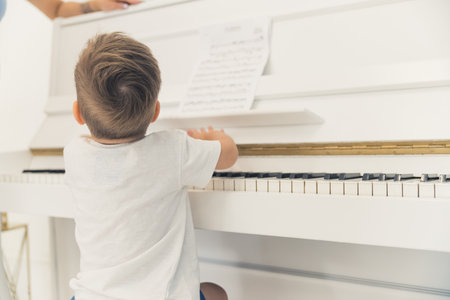 Little unrecognizable caucasian boy in a white t-shirt sitting in front of a beautiful ivory white piano. Music as a form of hobby for young kids. High quality photoの写真素材