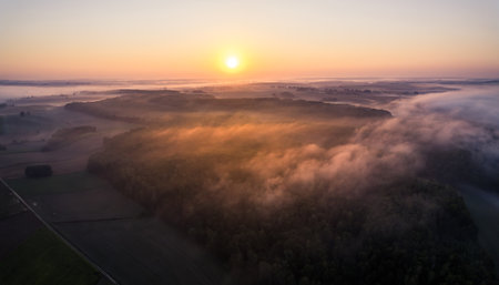 Bright orange rising sun over forest growing next to farm fields with fog all around. Orange glare. Morning view of Roztocze Poland. High quality photoの写真素材