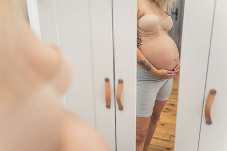 Pregnant caucasian woman with tattoos on her arms wearing gray biker shorts and nude strapless bra standing in front of a mirror and admiring her big belly. High quality photoの写真素材