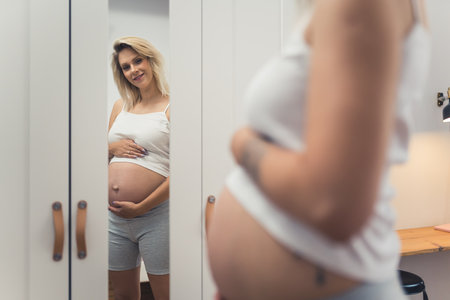 Caucasian young-adult blonde pregnant woman looking at her big belly in the wardrobe mirror. Indoor shot. Focus on the background. High quality photoの写真素材