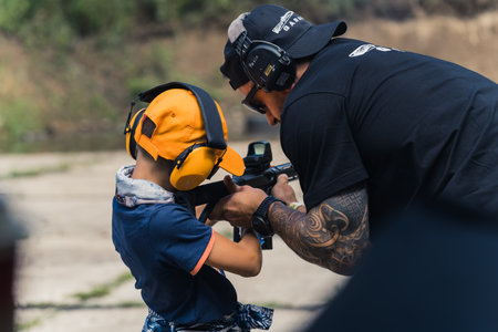 2022.08.07 Modlin, Poland - Male instructor showing child in safety headphones how to operate submachine gun. Gun training at firing range. Outdoor horizontal shot. High quality photoのeditorial素材