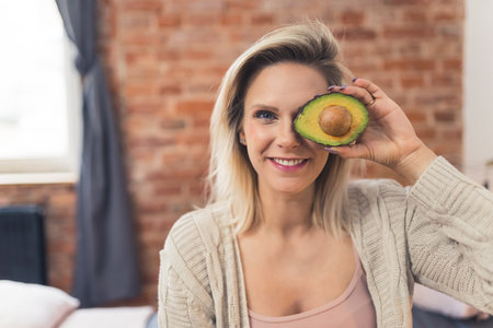 Happy smiling caucasian blond woman holding a half of avocado in front of her eye. Healthy...