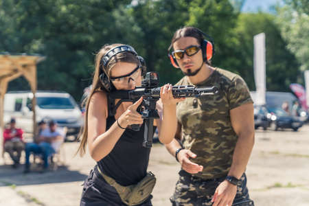 Bearded man in camo t-shirt watching woman aiming submachine gun. Safety headphones and goggles. Firearms training at firing range. Outdoor horizontal shot. High quality photoの写真素材