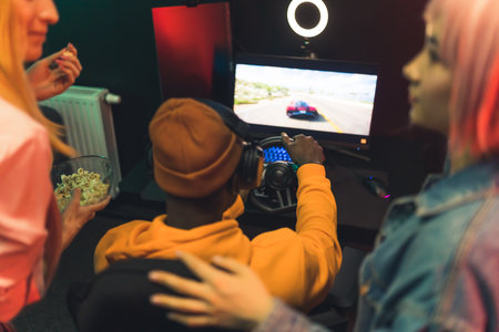 Two teenage girls talking watching teenage boy playing racing game using steering wheel. Gaming setup. Red lighting. . High quality photoの写真素材