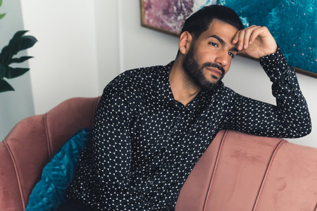 Hispanic young man with beard sitting on sofa holding hand up to forehead thinking and looking into distance. Indoor shot. High quality photoの写真素材