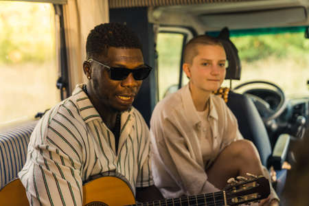 Multi-ethnic group of close friends on a summer road trip sitting inside their comfortable modern motorhome, playing the guitar and talking about different topics. High quality photoの写真素材