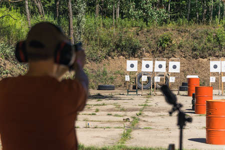 Rear view of man in safety headphones next to field glass practicing with hand gun on firing range. Target practice. Focus on background. Outdoor horizontal shot. High quality photoの写真素材