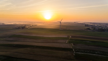 Colorful sunrise over panoramic farmfields with foggy horizon and huge wind turbine. Bright sun glare. Environmental awareness. Horizontal shot. High quality photoの写真素材