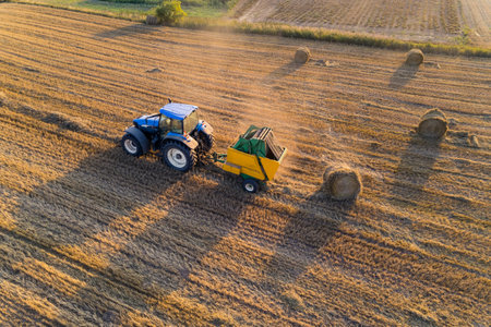 Golden field with bales of hay and blue tracktor stacking hay with green fields in the background. Agriculture. Horizontal shot. High quality photoの写真素材
