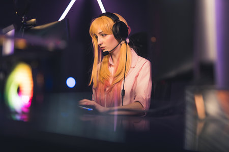 Young gamer girl wearing pink clothes and headset sitting in gaming chair playing an online game with determined look. Blurred foreground. High quality photoの写真素材