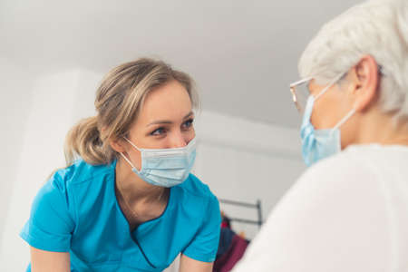 A beautiful young female doctor, wearing a blue uniform and a protective mask, smiling at her senior patient. High-quality photoの写真素材