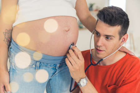 Excited soon-to-be father using stethoscope to hear the movement of his baby. Pregnant caucasian unrecognizable person showing her partner the sound of their child. High quality photoの写真素材