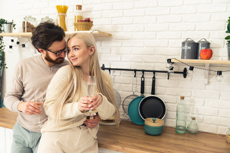 White couple brunette man and blonde woman in the kitchen hugging looking into each others eyes smiling and drinking champagne. Indooe shot. . High quality photoの写真素材