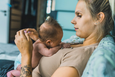 Skinny caucasian woman in her 30s lying down on bed and holding her little infant baby in a diaper on beige t-shirt. Blurred bedroom interior in the background. High quality photoの写真素材