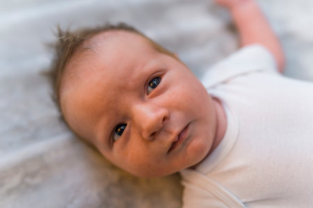 Close-up portrait of caucasian newborn baby boy lying on his bank on blanket on the floor wearing white t-shirt looking into camera. Horizontal shot. High quality photoの写真素材