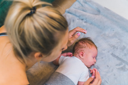 Caucasian blond woman leaning over her newborn baby boy lying on blanket on the floor next to couch. Caring mother. Responsibility. Horizontal indoor shot. High quality photoの写真素材
