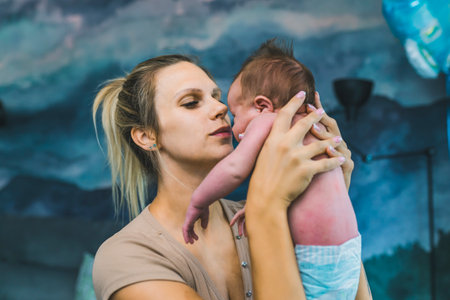 Caucasian happy loving mom holding her tiny newborn son in the air while standing in a blue-designed baby boy room at home. A woman enjoying playing with her child at an early age. High quality photoの写真素材