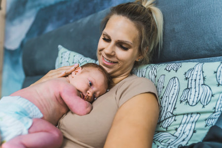Caucasian beautiful young mom relaxing in bed, smiling and looking at her newborn son peacefully laying in a fetal position on her chest and stomach. High quality photoの写真素材