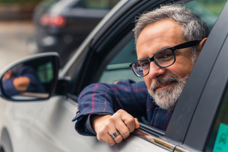Friendly caucasian man with gray hear, gray beard, and black glasses looking out of the driver's...