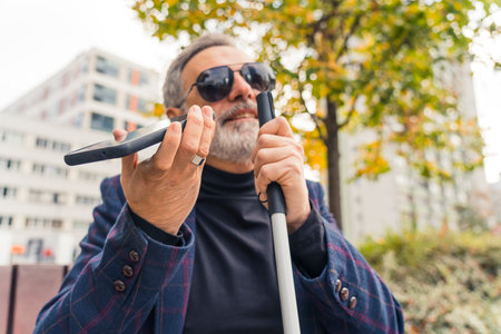 Elderly gray-haired caucasian entrepreneur with low vision in sunglasses sitting on bench, having a phone call using speaker mode, and holding white walking crane with one hand. Outdoor closeup shot. High quality photoの写真素材