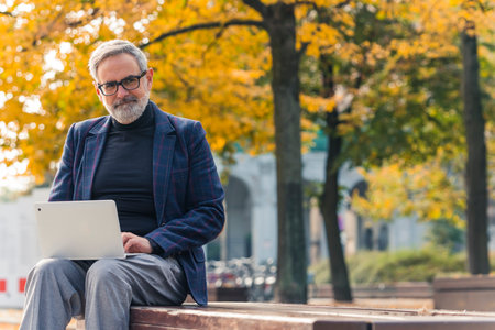 Elegant caucasian gray-haired man working remotely on his white laptop from park bench. Office break. Golden autumn. Sunny weather. Yellow and orange leaves. High quality photoの写真素材