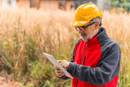 Professional adult caucasian architect looking through his glasses at construction plan on his tablet. Man in yellow hardhat and black-and-red fleece. Outdoor shot. High quality photoの写真素材
