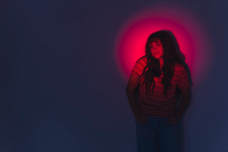 A young, beautiful girl with long dark hair leaning against the wall, smiling. Red circle light photoshoot. High quality photoの写真素材