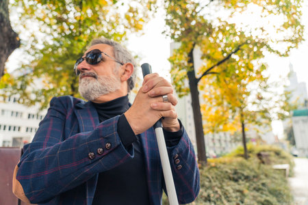 Blind bearded grey-haired mature man with dark sunglasses on holding a walking stick and sitting on a bench in the park. High quality photoの写真素材