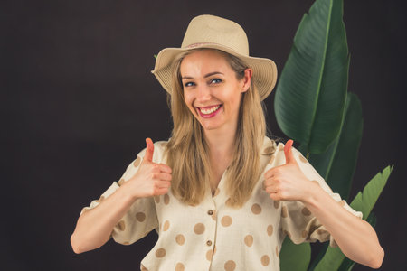 Smiling stylish blonde female with hat showing Good gesture with her fingers - black with plants...
