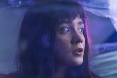 Close up face of a young, caucasian, beautiful girl with long dark hair and big worried eyes looking to the side. Photo through plastic wrap. Purple led lights. High quality photoの写真素材