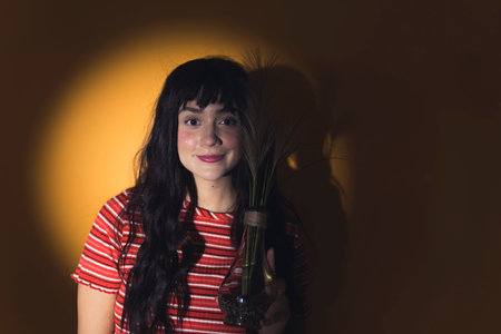 Young white woman with long dark hair standing in yellow spotlight looking at camera smiling holding vase with plant. Horizontal studio shot. High quality photoの写真素材