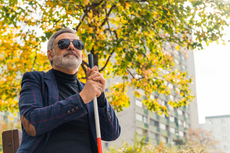 Blind bearded grey-haired mature man with dark eyeglasses on and a suit jacket holding a walking stick and sitting on a bench in the park. High quality photoの写真素材
