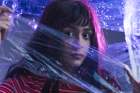 Caucasian teenage girl looking at camera with eyes wide open holding plastic wrap in front of her face. Dramatic expression. Artistic portrait. Horizontal shot. High quality photoの写真素材