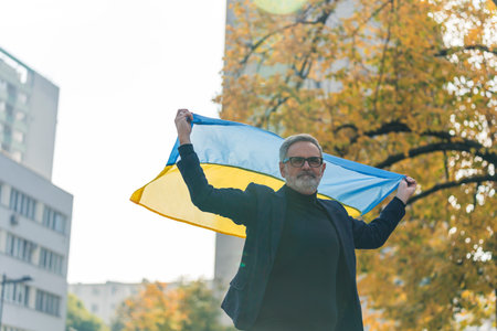 Confident, patriotic, bearded, grey-haired man in a blazer and a turtleneck with glasses on, holding the Ukrainian flag over his head. Blurred buildings and trees in the background. High quality photoの写真素材