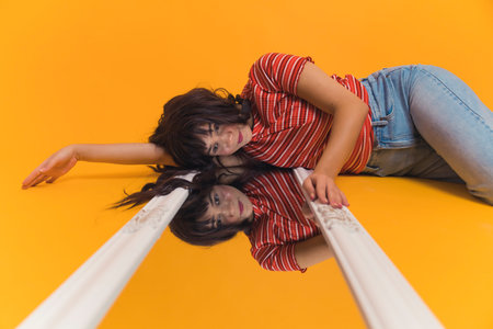 Young, bright, caucasian girl with long brown hair laying on the mirror on the floor. Girls reflection in the mirror, isolated orange background. High quality photoの写真素材