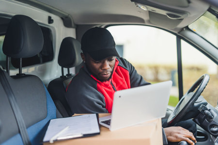 Handsome Black man in his 30s sitting inside a delivery van and checking parcel documents on silver laptop. Courier clothing. High quality photoの写真素材