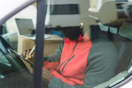 Unrecognizable Black adult man in black hat and red jacket working as a delivery person visible...