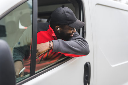 Black young adult delivery man sitting in driver seat of white van leaning out the window smiling looking away from camera. Horizontal shot. High quality photoの写真素材