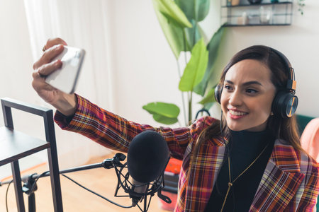 Young latin american female singer influencer recording song with professional setup holding up phone and smiling to take selfie. Horizontal indoor shot. High quality photoの写真素材