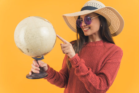 Young latin american woman wearing straw hat and sunglasses pointing at globe smiling. Travel destination. Tourism. Summer holidays. Yellow background horizontal studio shot. High quality photoの写真素材