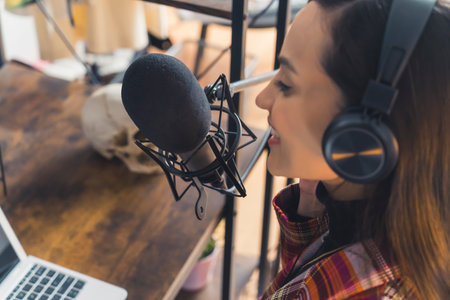 Latin american woman wearing headphones standing over desk singing into professional microphone looking at laptop recording song. Horizontal indoor shot. High quality photoの写真素材