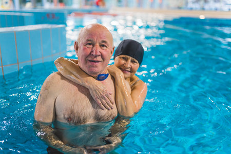 Lovely happy caucasian senior couple submerged in turquoise swimming pool water, looking at camera, and smiling. Active pensioners. Indoor portrait. High quality photoの写真素材