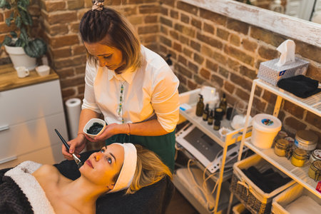 Cosmetologist applying black mask on the face of a beautiful blond woman at the spa, skincare...
