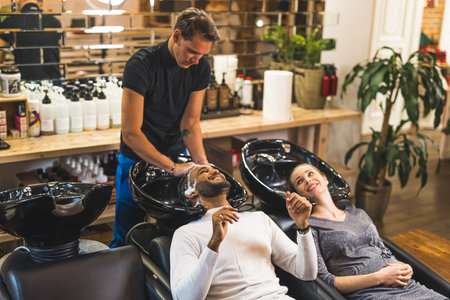 Happy indian male client having his hair washed with shampoo by hairdresser next to blonde female customer in a professional hair salon. High quality photoの写真素材