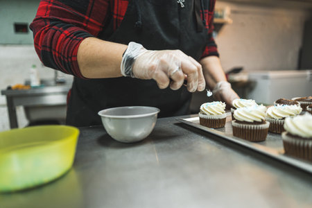 A baker wearing red plaid shirt, black apron ad transparent gloves decorating cupcakes with white cream placed on a baking tray in the kitchen. High quality photoの写真素材