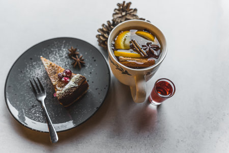 Dessert and a hot drink. A piece of cake decorated with powdered sugar and fruits placed on a black plate next to a white cup with a hot wintry drink and a small glass of red syrup. High quality photoの写真素材