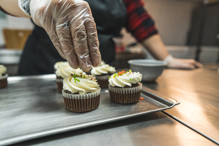 Close up shot of a person decorating cupcakes on a baking tray with colorful sprinkles. Hand in a...