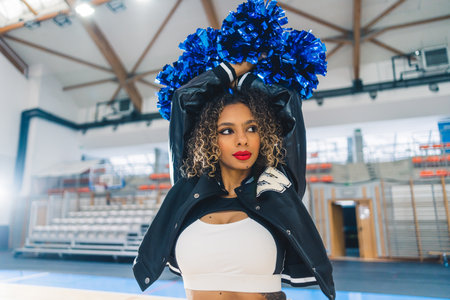 A brunette cheerleader with her arms lifted high and blue pom poms in her hands. High-quality photo