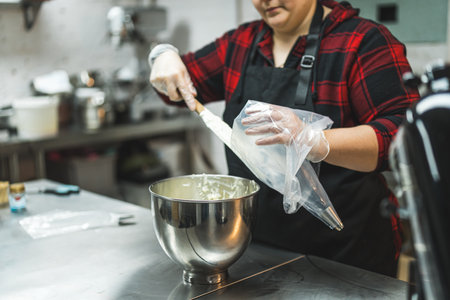 Female baker wearing gloves and black apron transfering buttercream frosting from silver bowl to piping bag. Professional baking process. Horizontal indor shot. High quality photoの写真素材