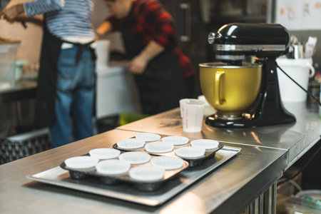Cupcake tray with paper liners next to stand mixer machine with two bakers in black apron working in...